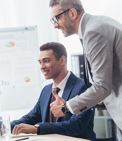 smiling businessman showing thumb up and looking at young colleague working with desk